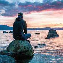 Man sitting on rocks in water, looking towards the sun low on the horizon, contemplating